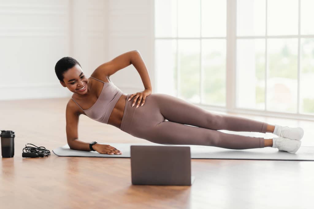Online Workout. Smiling Sporty Black Female Standing In Plank Position On Floor At Home Training, Looking At Laptop Watching Video Tutorial. Cheerful Lady Exercising. 