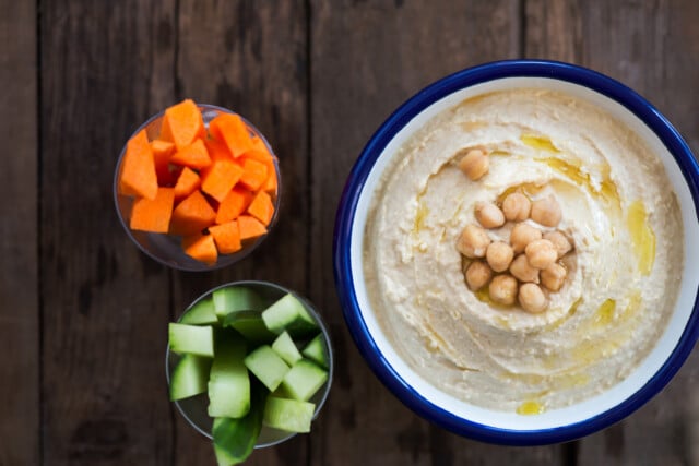 Classic Hummus made from Chickpeas in White Bowl, Carrot and Cucumber Sticks nearby, Wooden Rustic Background, top view