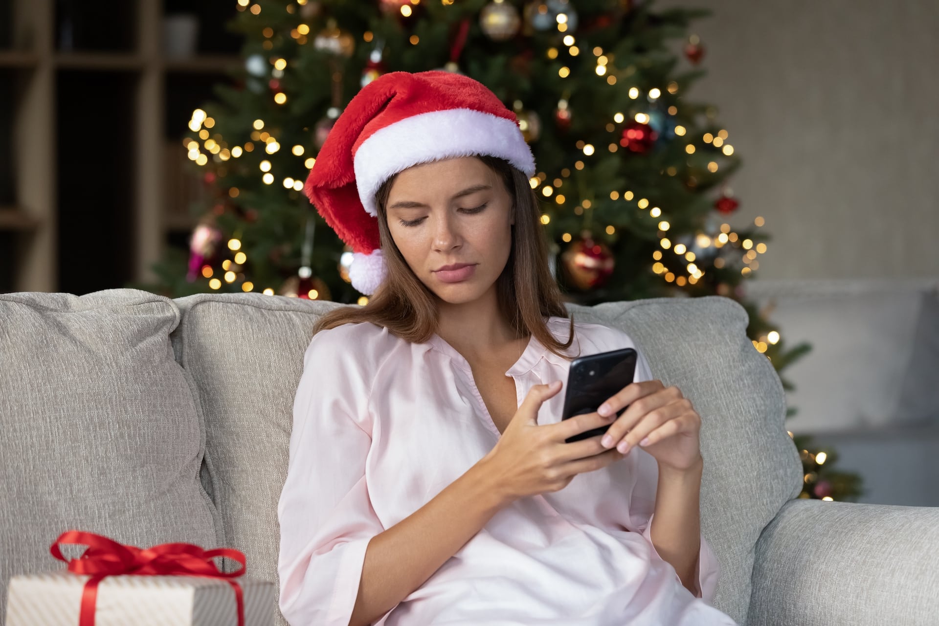 Sad thoughtful Xmas girl typing text message on smartphone, chatting online, resting on couch at gift box, Christmas tree. Woman with mobile phone feeling sad, frustrated, lonely at New Year night