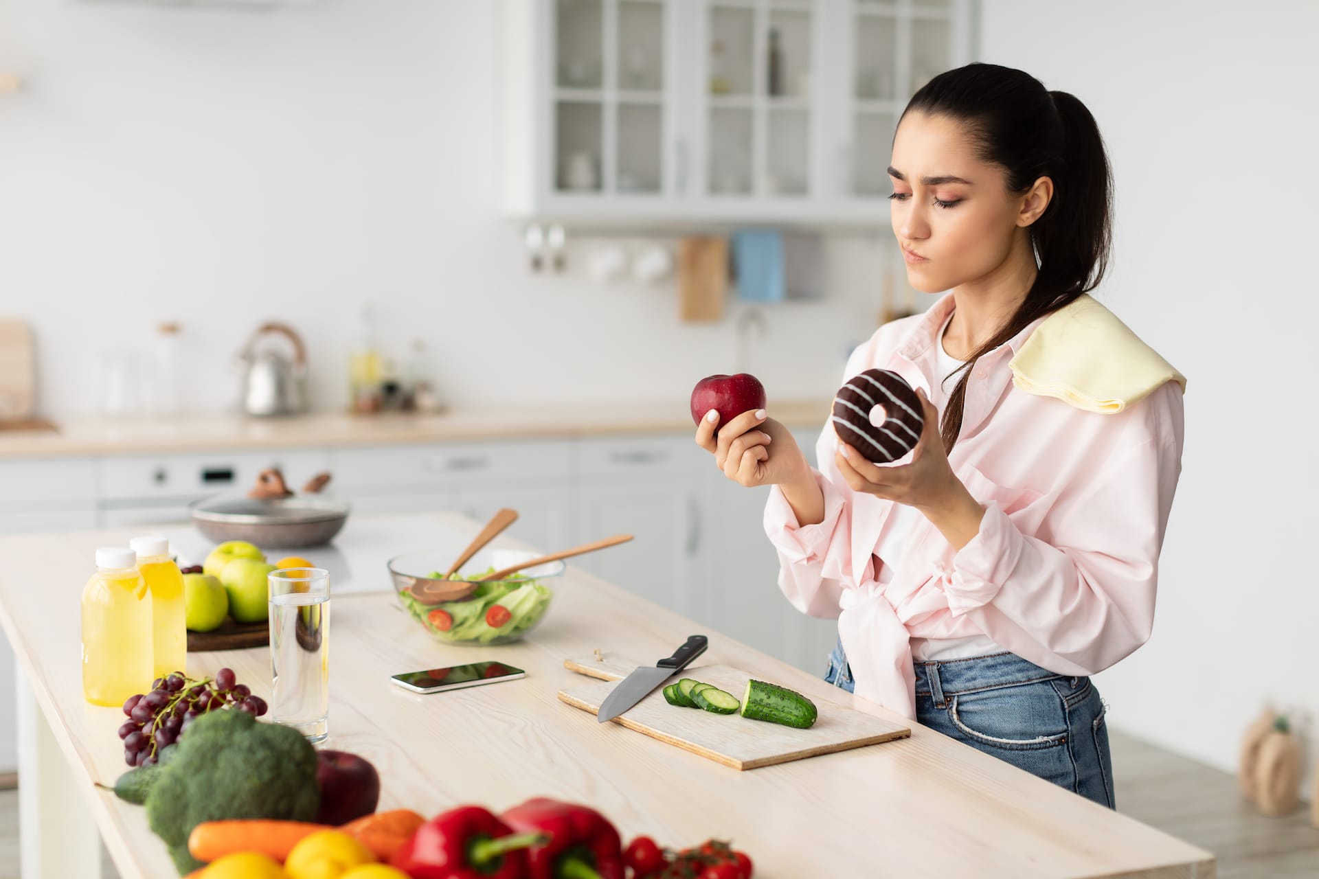Pretty pensive lady holding red apple and doughnut in her hands, looking at fruit. Young woman with sugar addiction wants to eat dessert but choosing good meal over unhealthy one