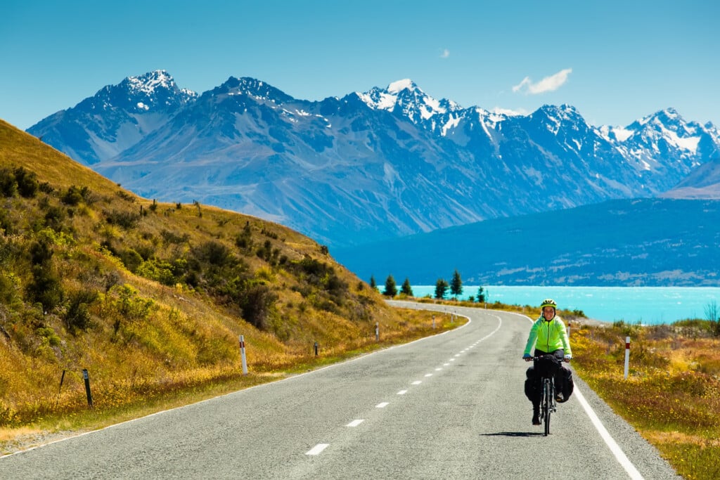 cyclist rides along the winding asphalt mountain road along Lake Pukaki view from Glentanner Park Centre near Mount Cook, on a background of blue sky with clouds, snowy Southern Alps.