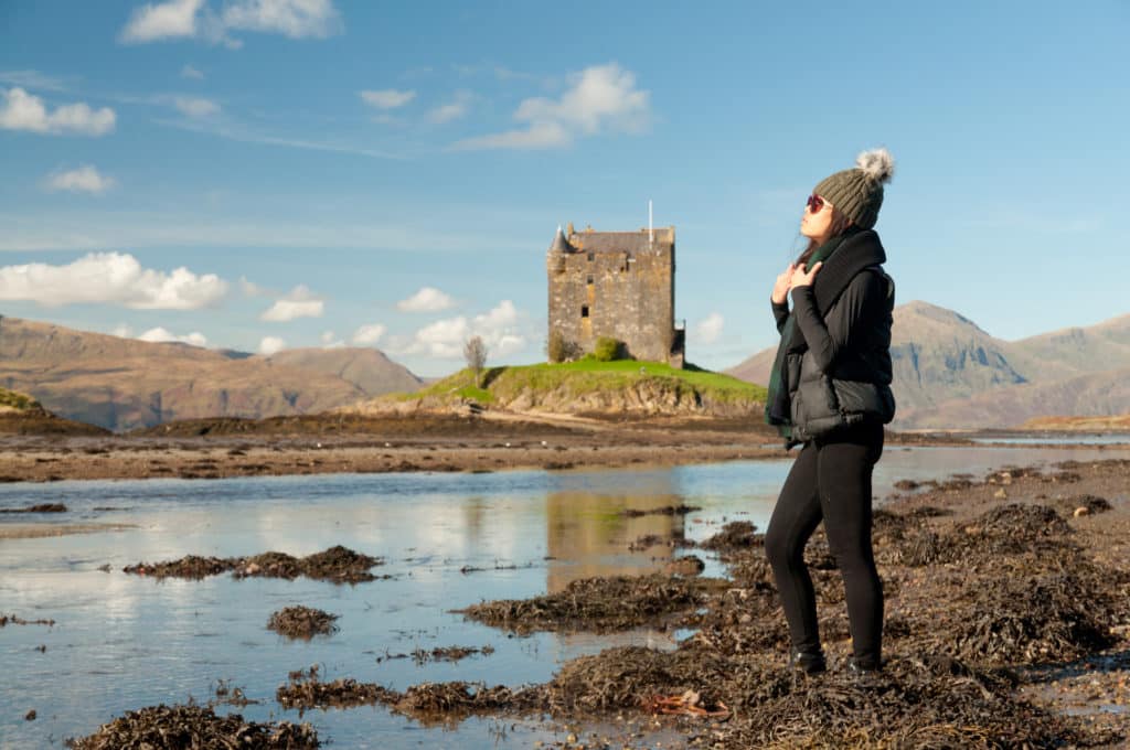 An Asian girl wearing a hat and sunglasses stands near the water surrounding the Stalker Castle near Port Appin, Argyll, Scotland, with the sun of her face as she looks away.