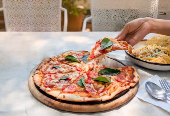 Hand is holding and pulling a slice of pizza margherita out of the tray,happy lunch time with friend in small garden. Foods on white tablecloth. Anabolic Fasting