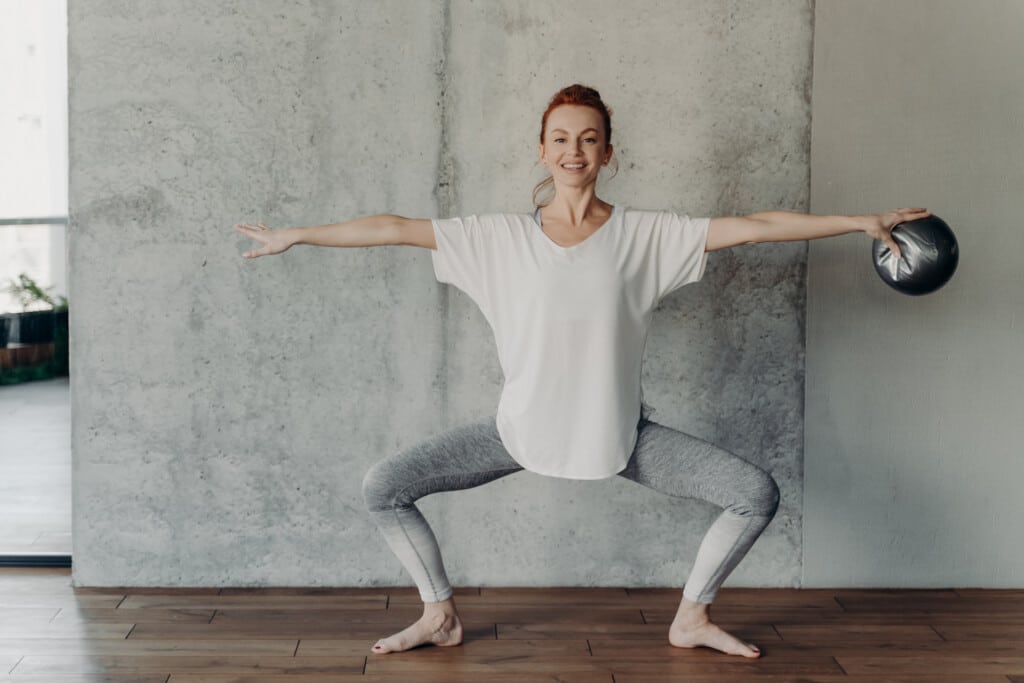 Happy fit redhaired woman sits in plie in second position with small fitball in one hand and does barre exercises or short leg workouts in fitness center ambience against grey concrete wall structure, dressed in sportswear