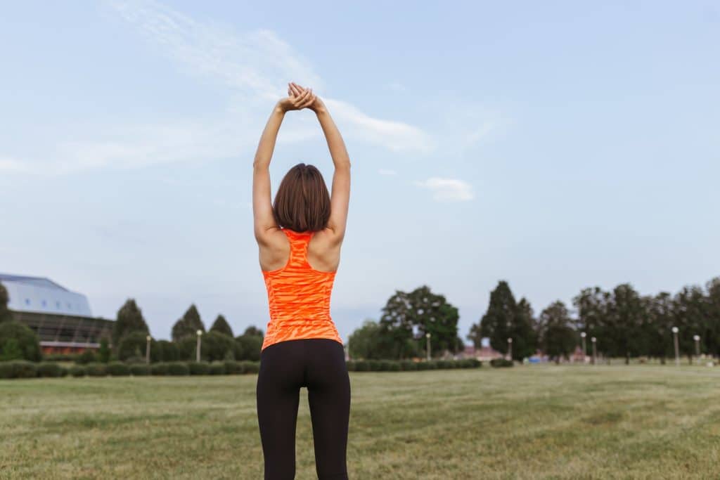 Back view of fit sportswoman in bright orange activewear stretching arms and warming up while training on field in summer. 